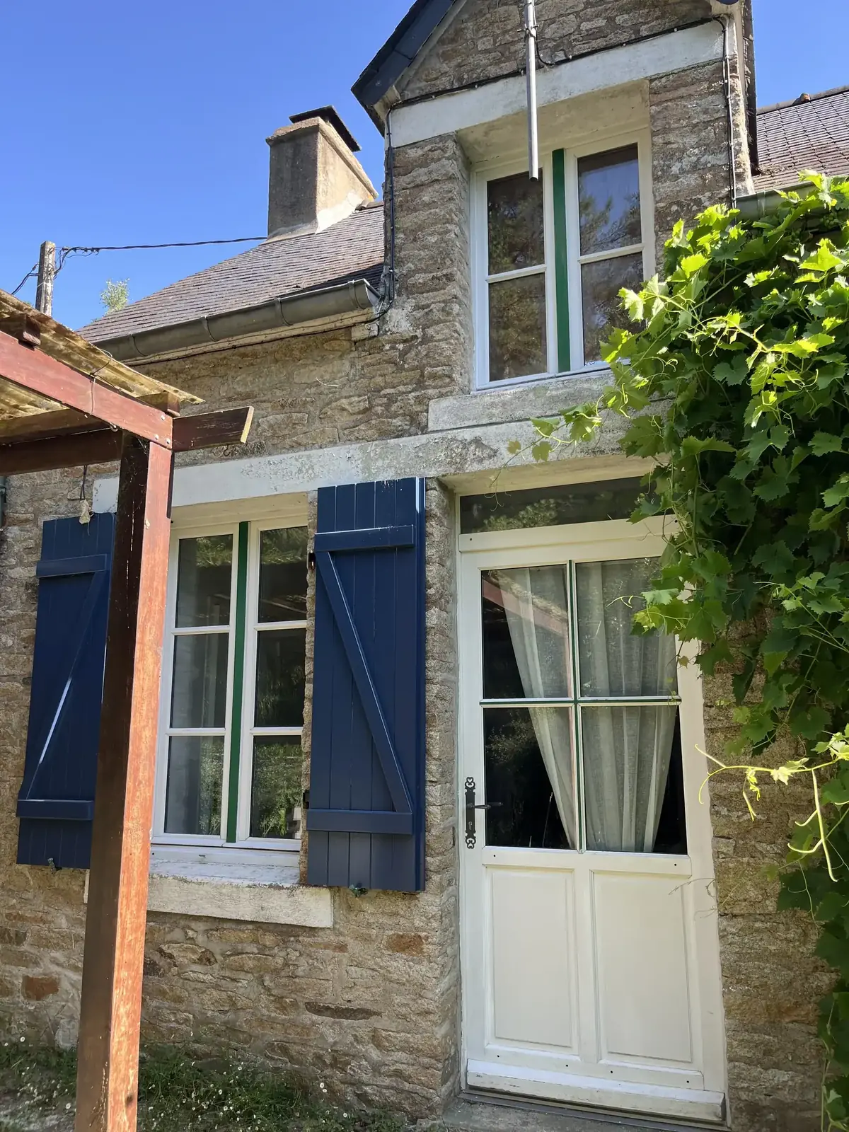 Entrée d’une longère de Peillac pendant le chantier, porte vitrée et fenêtre sous une pergola bois, volets bleus