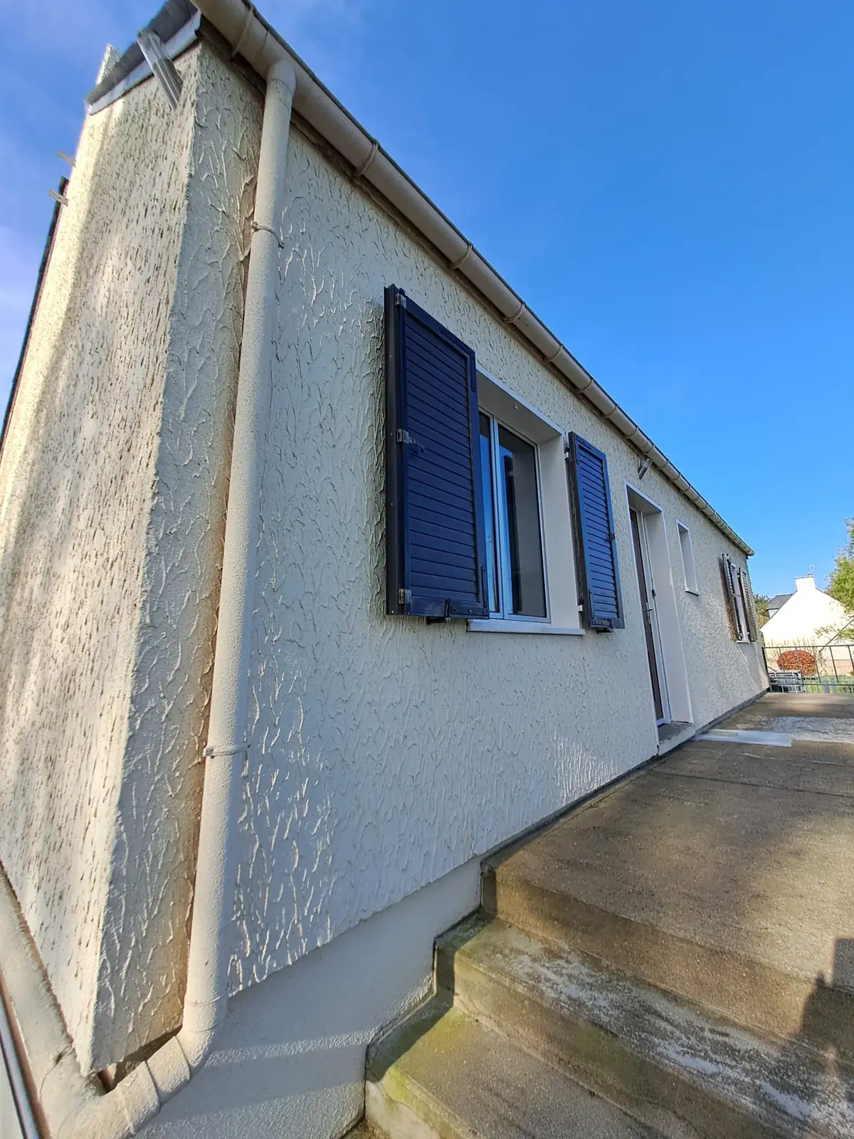 Façade blanche à enduit projeté d’une maison de Pleucadeuc, volets bleu soutenu et descente d’eau pluviale en pied de mur
