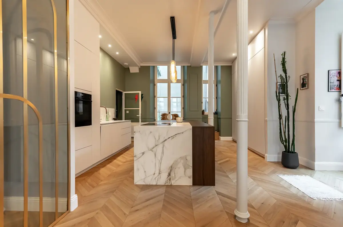 Cuisine d’un appartement ancien, verrière en arche dorée, îlot en marbre et parquet en chevrons